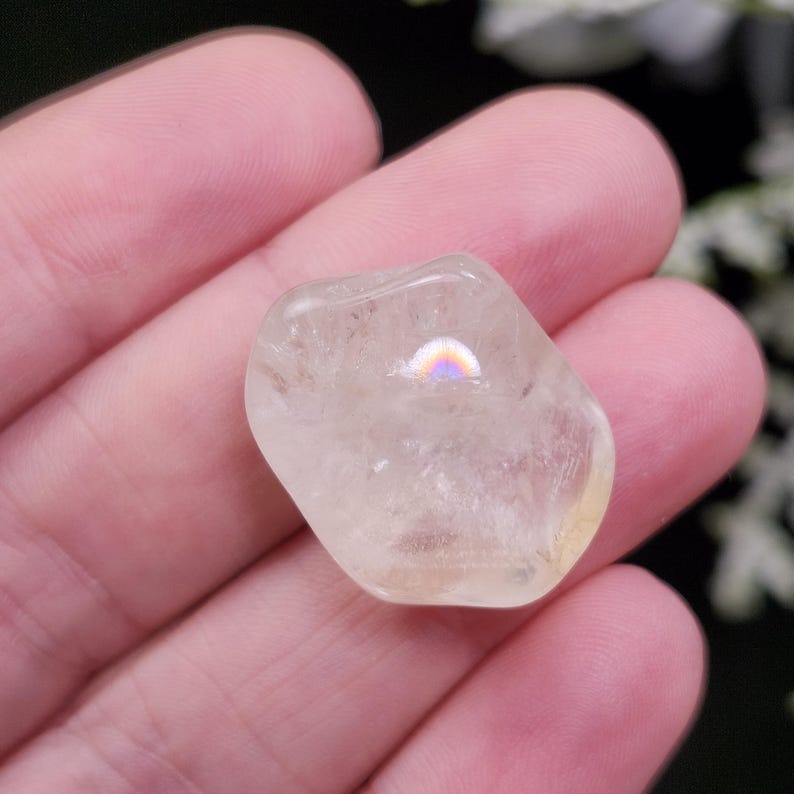 May include: A clear, polished crystal stone with a rounded shape. The stone has a subtle yellow hue and a rainbow-like iridescent effect on its surface. The crystal is held in a hand, showcasing its natural beauty and clarity.