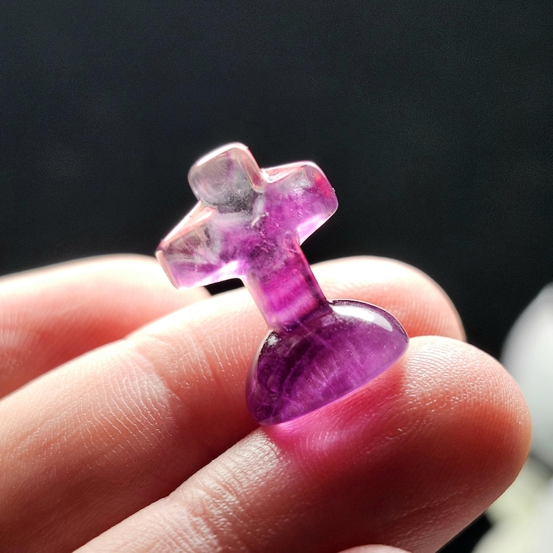 May include: A small, polished, cross-shaped gemstone in shades of purple and white. The cross has a rounded base and a smooth, translucent appearance, with visible internal patterns. The gemstone is held in a person's hand against a dark background.