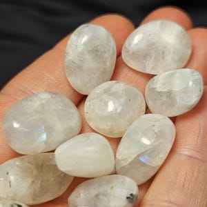 May include: A selection of polished moonstone gemstones, oval and rounded in shape. The stones are translucent with a milky white base and subtle iridescent flashes. The gemstones are held in a hand, against a dark background.