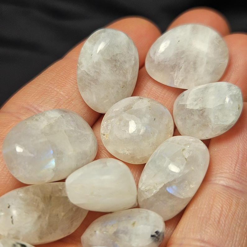 May include: A selection of polished moonstone gemstones, oval and rounded in shape. The stones are translucent with a milky white base and subtle iridescent flashes. The gemstones are held in a hand, against a dark background.