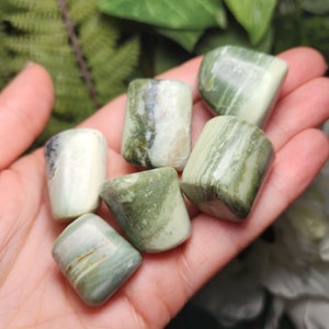 May include: A collection of tumbled green stones with varying shades and patterns. The stones are smooth and polished, with a mix of light and dark green hues. Some stones have white and gray streaks.