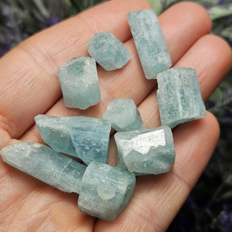 K&ouml;nnte beinhalten: Eine Sammlung roher Aquamarinkristalle in verschiedenen Formen und Gr&ouml;&szlig;en. Die Kristalle haben eine blassblau-gr&uuml;ne Farbe mit sichtbaren nat&uuml;rlichen Texturen und Facetten. Die Kristalle werden in einer Hand gehalten, was ihre nat&uuml;rliche Sch&ouml;nheit zeigt.