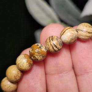 May include: Close-up of a beaded bracelet featuring round, natural stone beads. The beads are primarily tan with brown and black veining, creating a unique pattern. The bracelet is held in a hand, showcasing the texture and color variations.