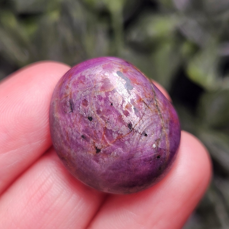 May include: A polished, oval-shaped gemstone in shades of deep pink and purple. The stone has a smooth, reflective surface with subtle variations in color and small black inclusions. The gemstone is held in a hand, with a blurred green background.