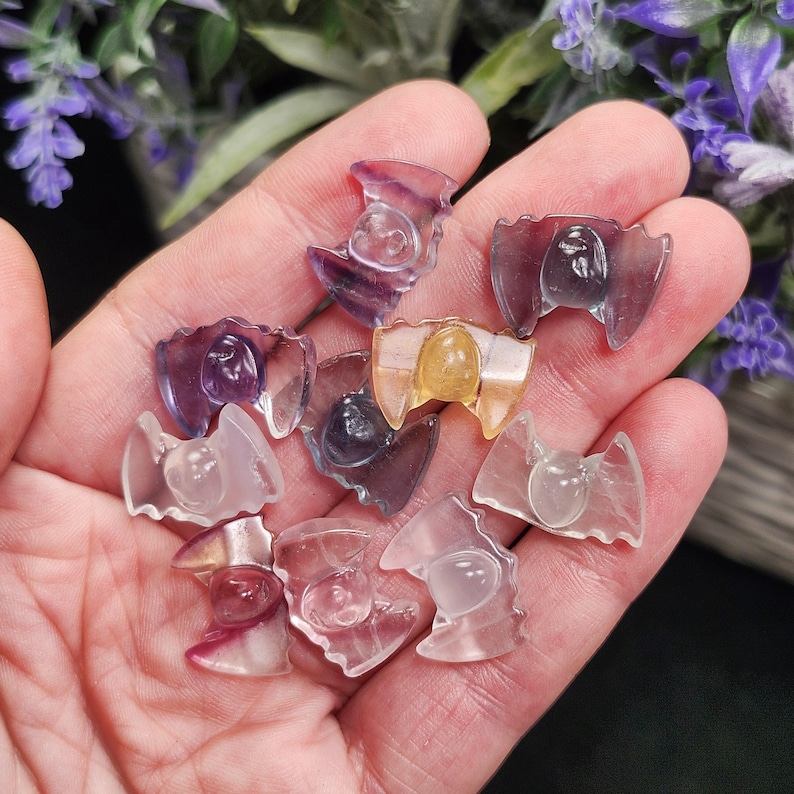 May include: A collection of small, carved crystal bat figurines in various colors, including purple, yellow, and clear. The bats have detailed wings and a rounded body. The figurines are held in a hand, with a blurred background of purple flowers.