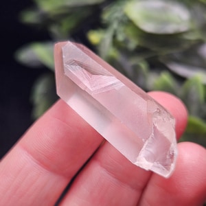May include: A clear, elongated quartz crystal point with a distinct phantom inclusion visible inside. The crystal has a smooth, faceted surface with a pointed tip and a slightly irregular, fractured base, showcasing natural internal geometric patterns.