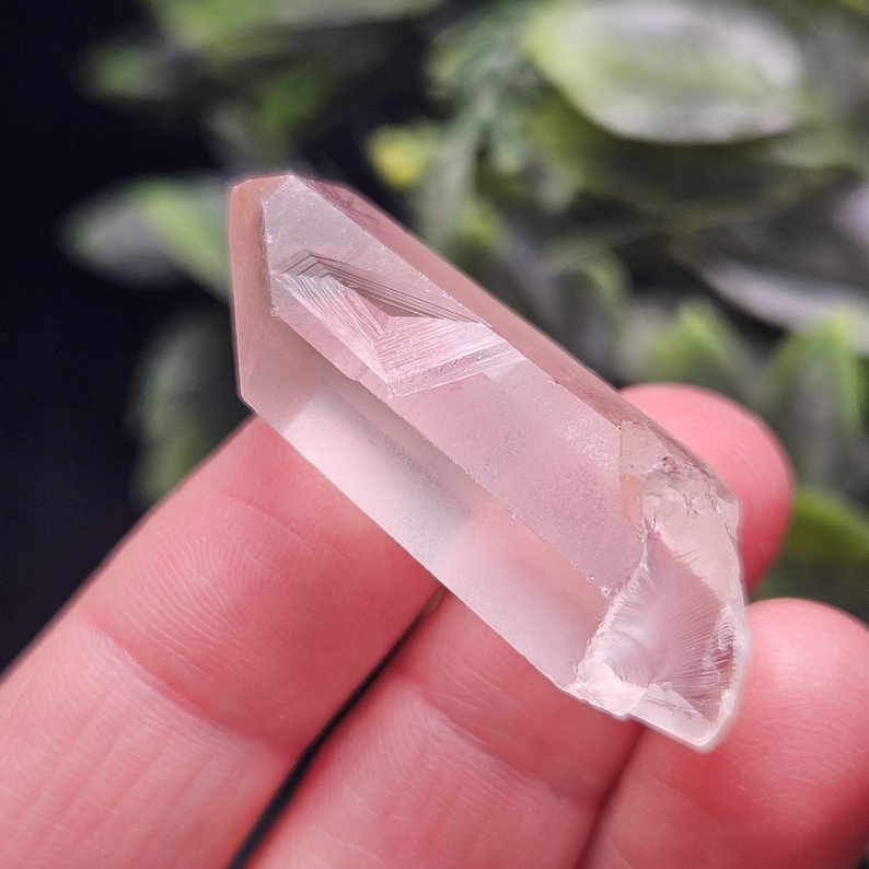 May include: A clear, elongated quartz crystal point with a distinct phantom inclusion visible inside. The crystal has a smooth, faceted surface with a pointed tip and a slightly irregular, fractured base, showcasing natural internal geometric patterns.
