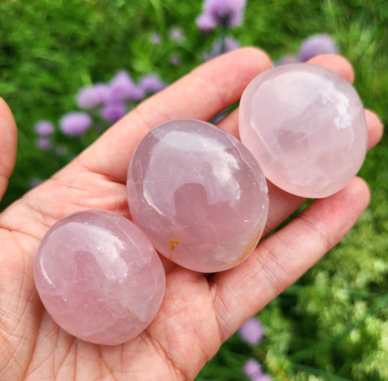K&ouml;nnte beinhalten: Drei glatte, polierte Rosenquarzsteine mit einem durchscheinenden, blassrosa Farbton. Jeder Stein hat eine abgerundete, ovale Form mit einer gl&auml;nzenden, reflektierenden Oberfl&auml;che. Die Steine variieren leicht in der Gr&ouml;&szlig;e und weisen nat&uuml;rliche Einschl&uuml;sse auf.