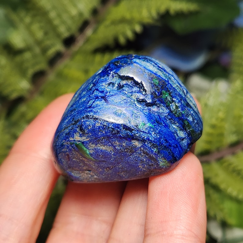 May include: A polished, rounded stone with a deep blue hue, accented by streaks of green and lighter blue. The stone's surface has a textured appearance, with natural variations in color and pattern. The stone is held in a hand, with a blurred green background.