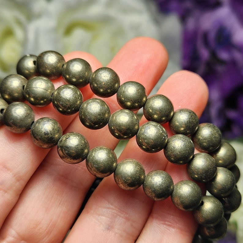 May include: Three strands of pyrite gemstone beads, each approximately 8mm in diameter, are displayed. The beads have a metallic, golden-grey colour and a smooth, rounded shape. The bracelets are held in a hand, with a blurred floral background.