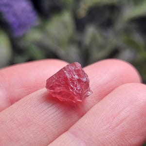May include: A small, raw, translucent gemstone with a deep pinkish-red hue. The stone features an irregular, multifaceted surface with sharp, jagged edges and a crystalline structure that catches the light, displaying varying shades of red.
