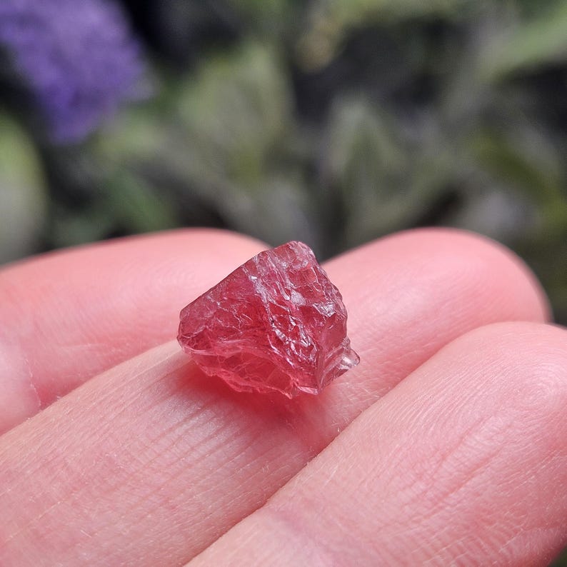 May include: A small, raw, translucent gemstone with a deep pinkish-red hue. The stone features an irregular, multifaceted surface with sharp, jagged edges and a crystalline structure that catches the light, displaying varying shades of red.