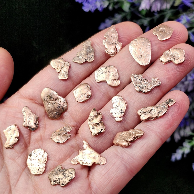 May include: A collection of small, irregularly shaped gold nuggets. The nuggets vary in size and texture, with a mix of smooth and rough surfaces. The gold nuggets are displayed on a hand, with a blurred background of purple flowers.