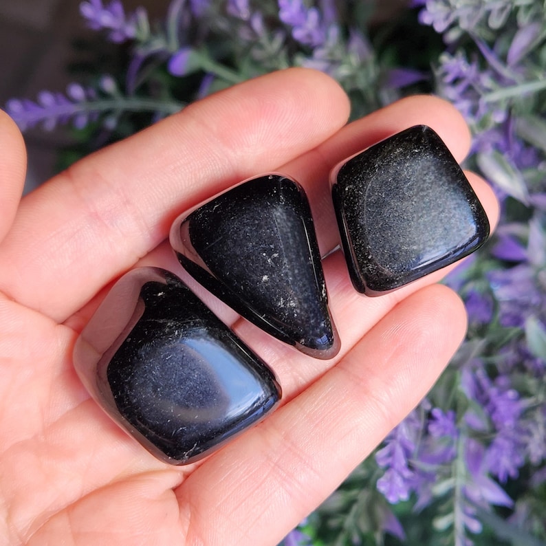 May include: Three polished black stones with a smooth, reflective surface and subtle metallic flecks. These stones are irregular in shape, featuring rounded edges and a dark, glossy appearance that catches the light, showcasing a deep, solid black color.