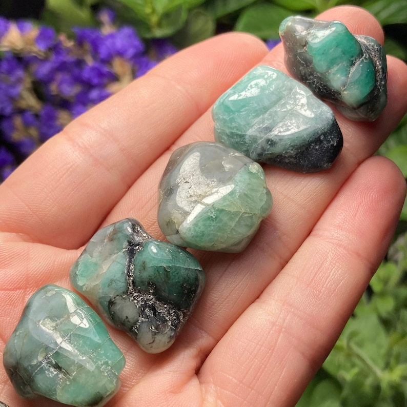 May include: Five raw emerald stones in varying shades of green and grey are displayed in a hand. The gemstones have a rough, natural texture and are irregularly shaped. The background features blurred green foliage and purple flowers.