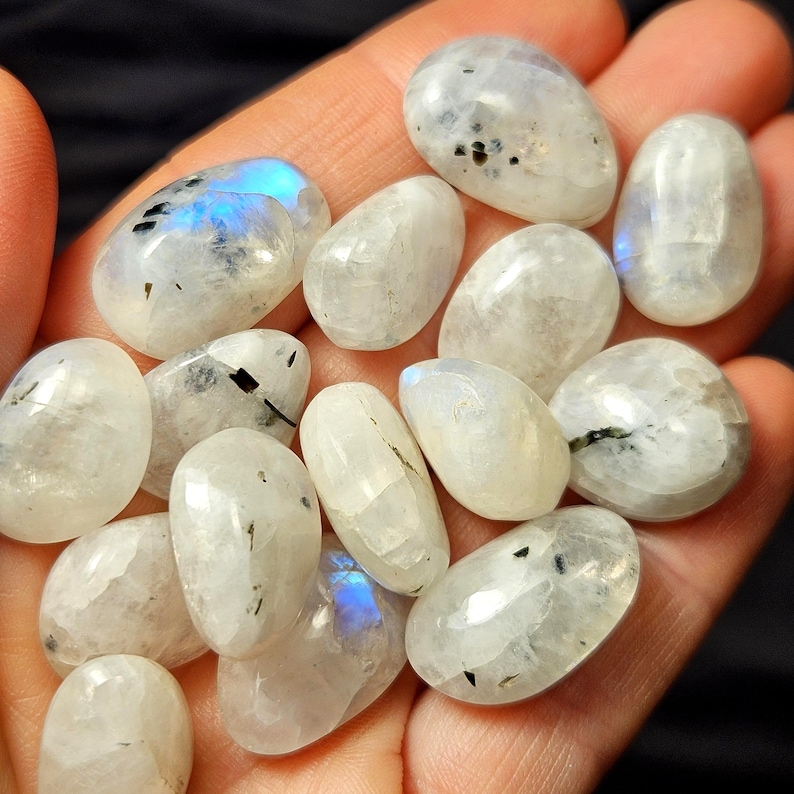 May include: A selection of polished moonstone gemstones. The stones are oval-shaped, with a milky white base colour and iridescent blue flashes. Some stones have small black inclusions. The gemstones are held in a hand.