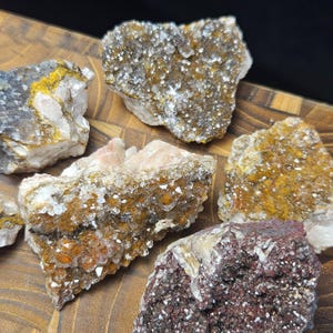 May include: A collection of raw, uncut crystals in various colors, including white, yellow, orange, and deep red. The crystals have a rough, textured surface and are displayed on a wooden cutting board.