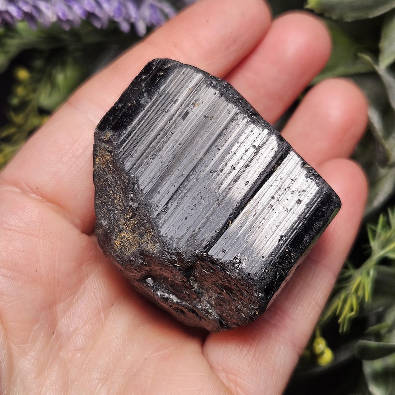 Peut inclure: Un cristal de tourmaline noire brute pr&eacute;sentant une couleur noire profonde avec une surface brillante et r&eacute;fl&eacute;chissante. La pierre affiche des stries verticales marqu&eacute;es sur toute sa longueur, avec une base rugueuse et textur&eacute;e.