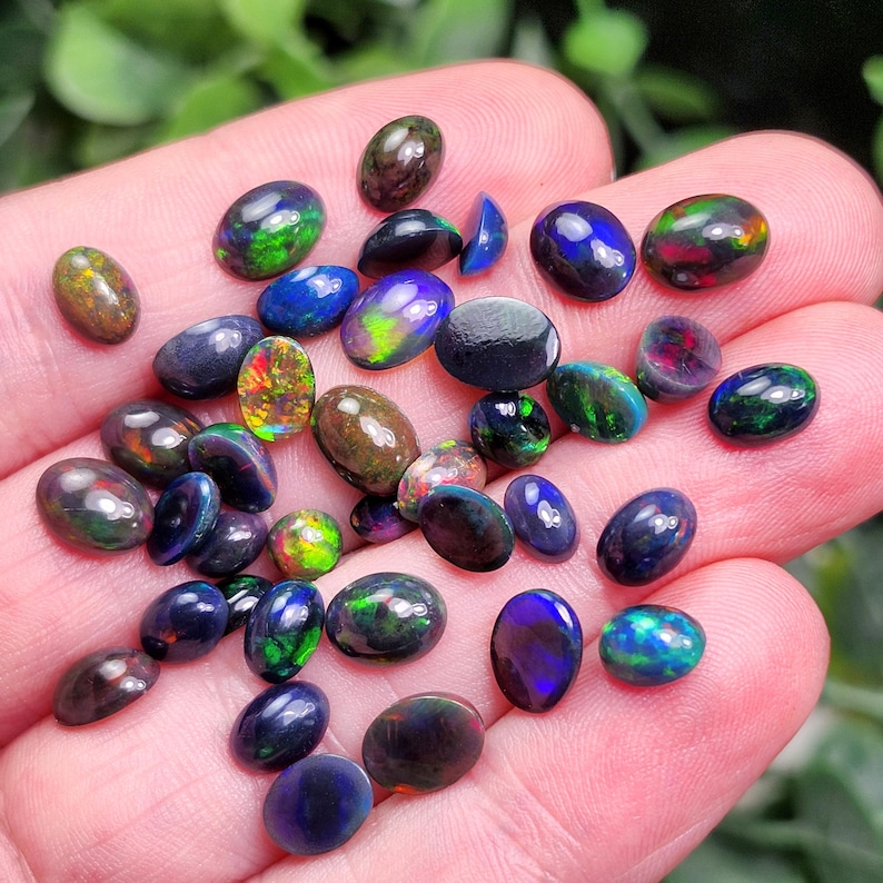 May include: A collection of oval-shaped opals in various colors, including blue, green, red, and brown. The opals are displayed in a hand, showcasing their iridescent qualities. The gemstones vary in size and shape, with a polished, smooth surface.
