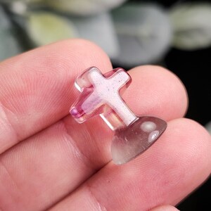 May include: A small, translucent cross-shaped object with a rounded base. The cross is primarily pink with a hint of purple, and the base is a gradient of purple to clear. The object is held in a hand.