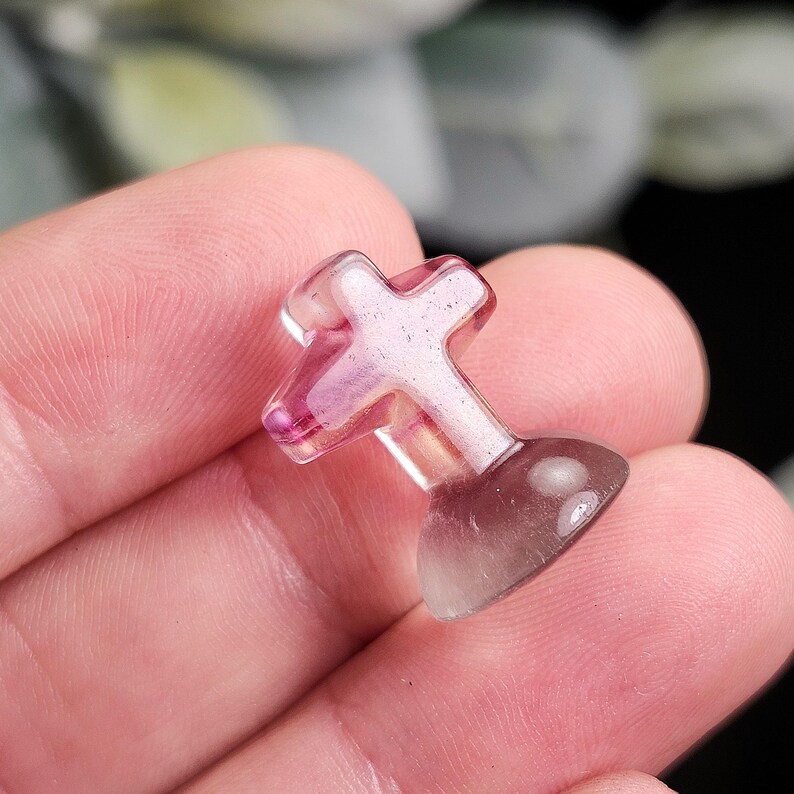May include: A small, translucent cross-shaped object with a rounded base. The cross is primarily pink with a hint of purple, and the base is a gradient of purple to clear. The object is held in a hand.