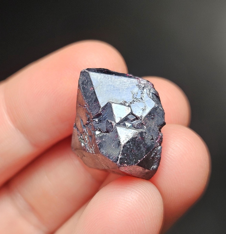 Puede incluir: Un esp&eacute;cimen mineral oscuro y facetado con un brillo profundo y reflectante. El cristal exhibe una estructura geom&eacute;trica compleja, con bordes afilados y un color oscuro, casi negro. La superficie refleja la luz.