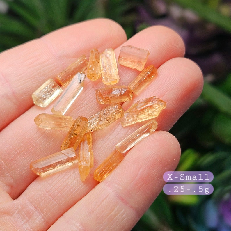 May include: A collection of small, translucent, orange-toned crystal shards with elongated, prismatic shapes. A purple graphic overlay in the lower right corner displays the text X-Small .25-.5g, indicating the weight range in grams.