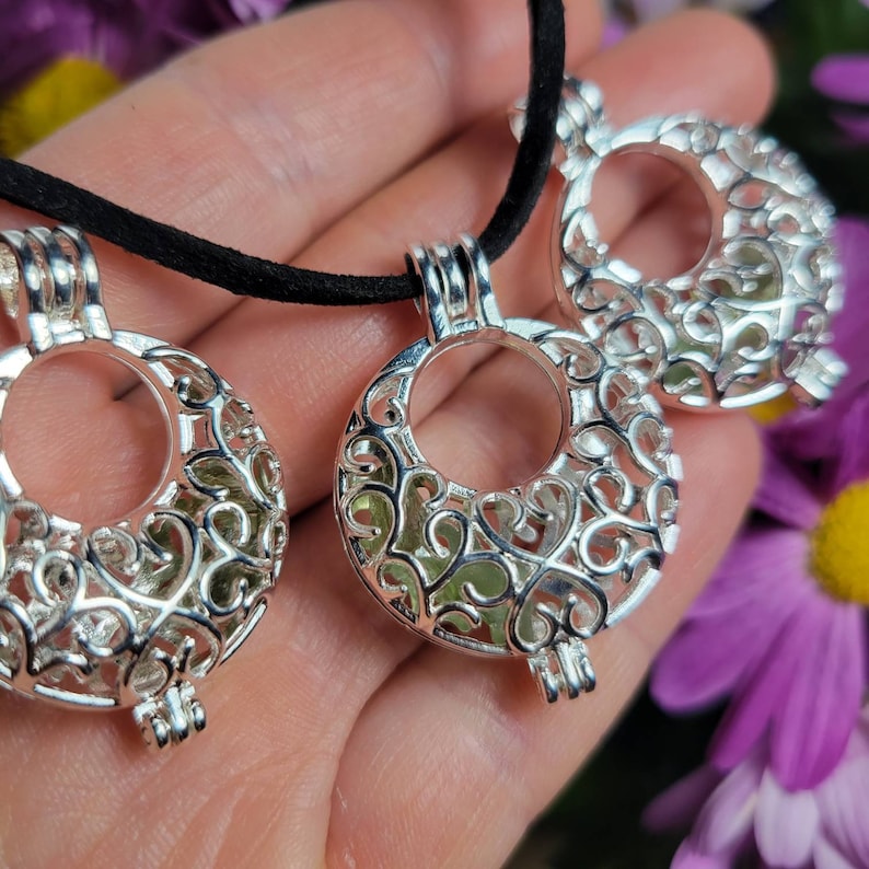 May include: A silver necklace with three pendant charms. Each charm is a filigree cage with a round opening. The cages are filled with green crystals.