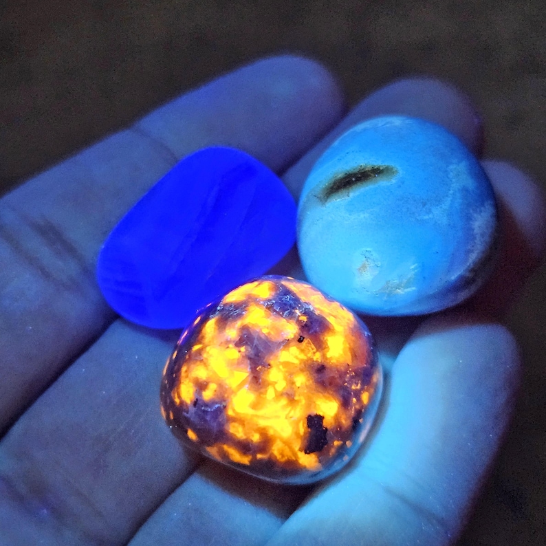 May include: Three glowing gemstones in a hand. One is a vibrant blue, another is a mix of orange and purple, and the third is a light blue with a dark streak. The stones are smooth and rounded, with a bright, almost neon, appearance.