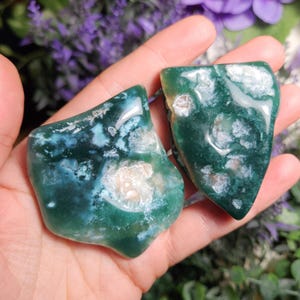 May include: Two green moss agate stones with white and brown inclusions. The stones are irregularly shaped and have a smooth, polished surface.