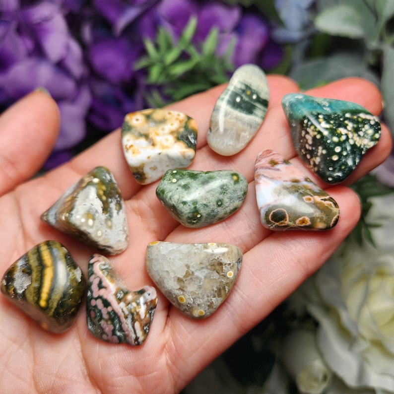 May include: A collection of nine ocean jasper tumbled stones in various shades of green, brown, white, and pink with white and brown spots. The stones are smooth and polished.