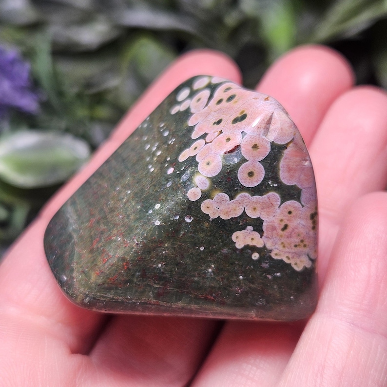 Puede incluir: Una piedra de jaspe oce&aacute;nico pulida de forma triangular que presenta una base de color verde bosque oscuro con distintos patrones orbiculares circulares de color rosa y crema dispersos por la superficie, con una textura suave, brillante y reflectante.