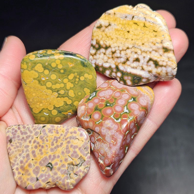 May include: Four polished gemstones with unique patterns and colors. One is primarily yellow with green spots, another is a mix of red and pink with white lines, and two others feature yellow, purple, and brown hues. The stones are held in a hand.