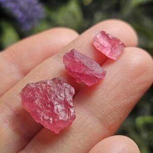 May include: Three raw, unpolished pink tourmaline gemstones with irregular, jagged shapes and translucent, crystalline textures. The stones vary in size, displaying deep magenta and soft rose hues with natural facets and internal fractures.
