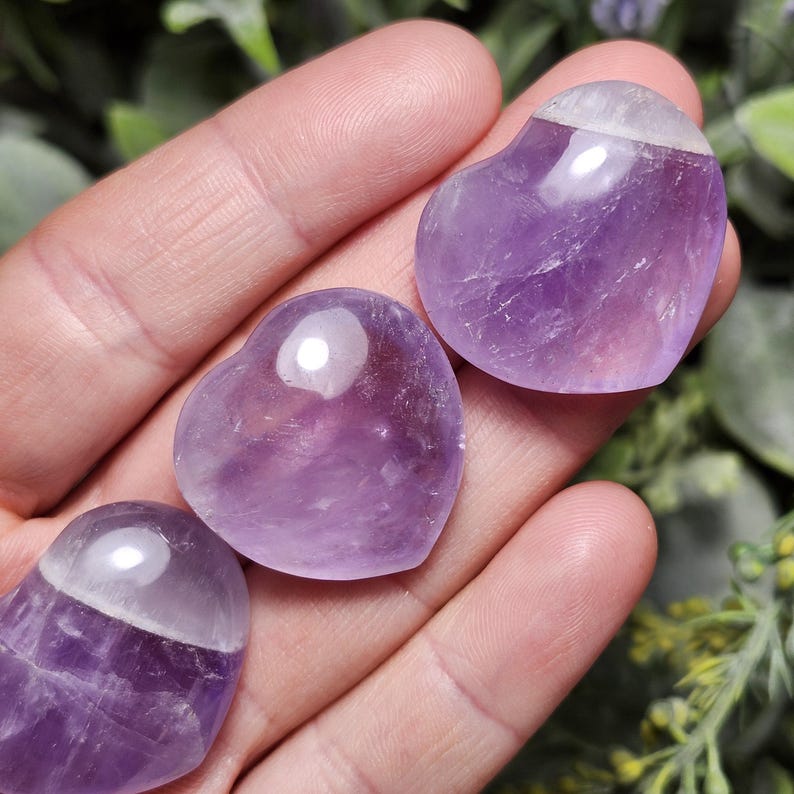 Puede incluir: Tres cristales de amatista en forma de coraz&oacute;n, pulidos y lisos, con un tono p&uacute;rpura claro. Los cristales est&aacute;n dispuestos en una mano, con el cristal m&aacute;s grande en la parte superior.