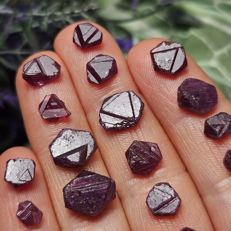 K&ouml;nnte beinhalten: Eine Sammlung von 13 tiefvioletten Rubinkristallen, jeder mit einer einzigartigen, facettierten Form. Die Kristalle sind auf einer Hand angeordnet und zeigen ihre komplizierten Details.