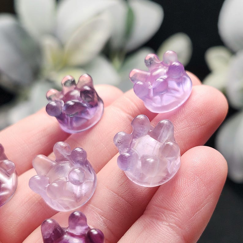 May include: Several small, translucent, purple-hued crystal carvings shaped like hands. The carvings have rounded fingers and a smooth, polished appearance. The crystals vary in shades of purple, from light lavender to deep violet.