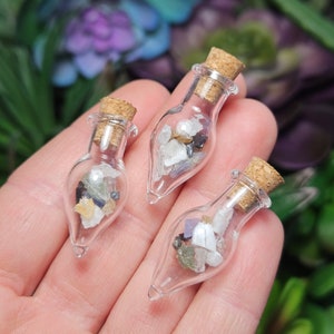 May include: Three small glass bottles with cork stoppers filled with various colorful crystals and stones. The bottles are clear and have a teardrop shape.