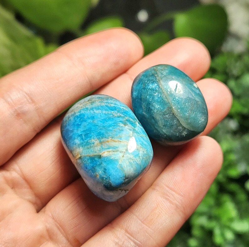 May include: Two polished, oval-shaped blue gemstones with varying shades of turquoise and streaks of brown. The stones are smooth and reflective, with a natural, organic appearance. The gemstones are held in a hand.