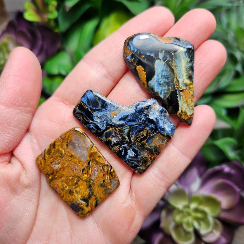 May include: Three polished gemstones with irregular shapes. One stone is primarily golden-brown with dark streaks, while the other two feature a mix of deep blue, black, and golden-brown swirling patterns, showcasing a chatoyant, fibrous texture.