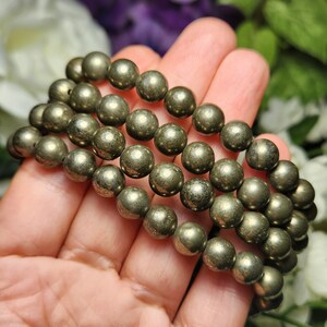 May include: A pyrite bracelet with multiple strands of spherical, metallic gold-coloured beads. The beads have a textured surface and are strung together. The bracelet is displayed against a soft, blurred floral background.
