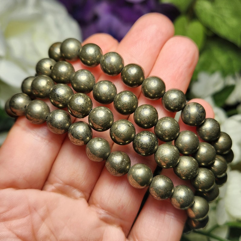 May include: A pyrite bracelet with multiple strands of spherical, metallic gold-coloured beads. The beads have a textured surface and are strung together. The bracelet is displayed against a soft, blurred floral background.