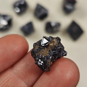 Puede incluir: Primer plano de un esp&eacute;cimen mineral cristalino oscuro. La atenci&oacute;n se centra en un grupo de cristales oscuros de aspecto met&aacute;lico con un cristal triangular prominente y reflectante. Otros espec&iacute;menes similares est&aacute;n en el fondo.