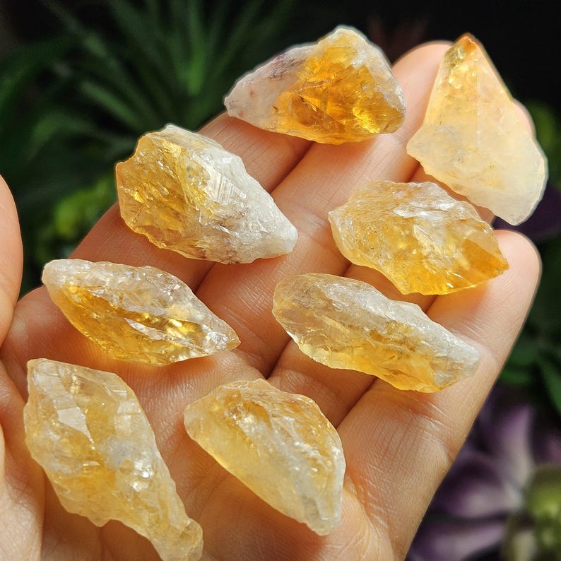 May include: A collection of eight raw citrine crystal stones with translucent yellow and orange hues. Each stone features jagged, irregular edges and a natural crystalline texture with varying degrees of clarity and internal facets.