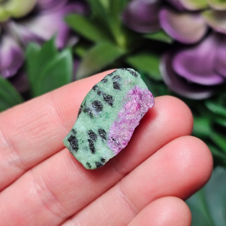 Pu&ograve; includere: Una gemma grezza di rubino zoisite. La pietra presenta una base verde con macchie nere e una sezione di rubino rosa vibrante. La consistenza ruvida e naturale della pietra &egrave; visibile, mostrando la sua forma organica. La gemma &egrave; tenuta in una mano.