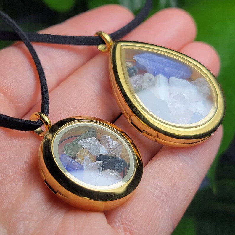 May include: Two gold locket necklaces with clear glass windows. The larger locket is teardrop shaped and the smaller locket is round. Both lockets are filled with colorful crystals and stones.