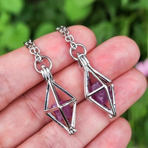 May include: Two silver necklaces with a geometric cage pendant. Each cage holds a small, purple crystal.