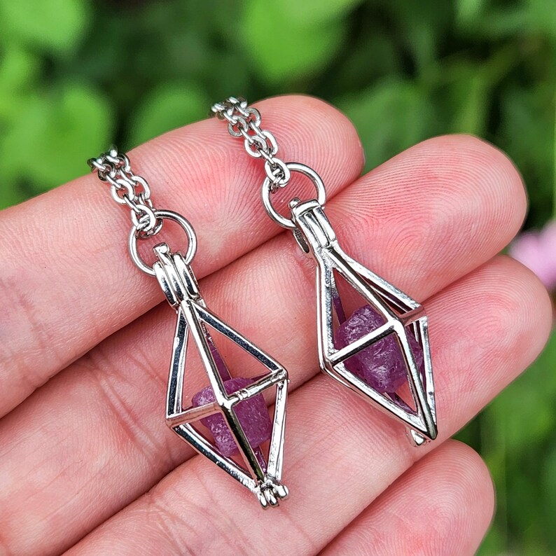 May include: Two silver necklaces with a geometric cage pendant. Each cage holds a small, purple crystal.