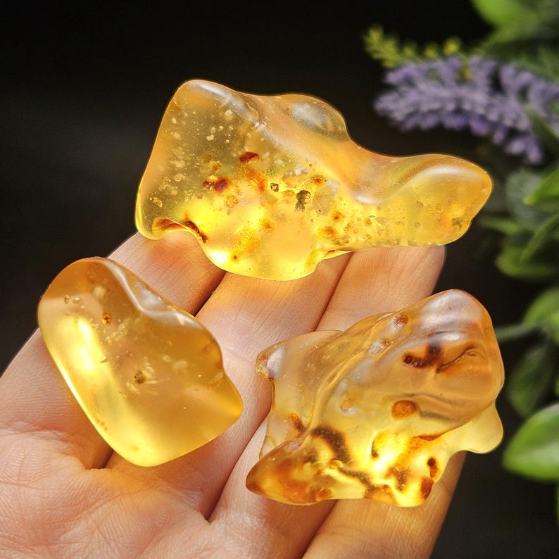 May include: Three polished, translucent amber stones with irregular, organic shapes. The stones exhibit a warm, golden-yellow hue with visible internal inclusions, dark brown organic flecks, and a smooth, glossy surface texture throughout.