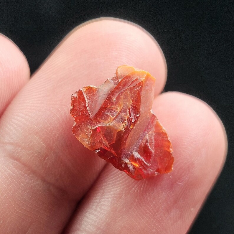 May include: A rough-cut, translucent, orange-red gemstone with a crystalline structure. The gem has a fiery appearance, with internal light reflections. The stone is held between fingers against a dark background, highlighting its color and texture.